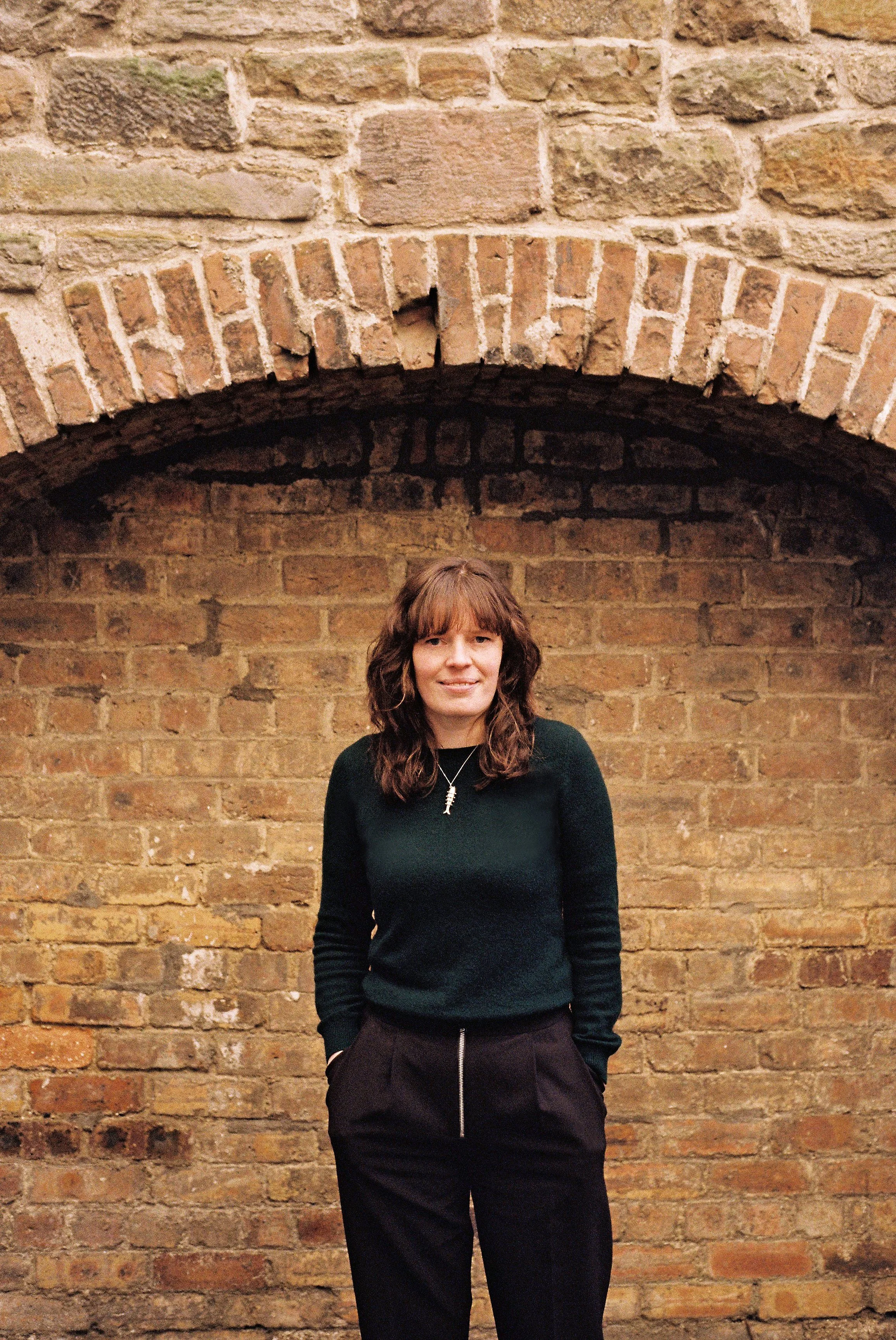 A woman with shoulder-length brown hair standing in front of a brick wall with a large brick arch above her head. She is wearing a dark green sweater and black pants, and smiling at the camera.