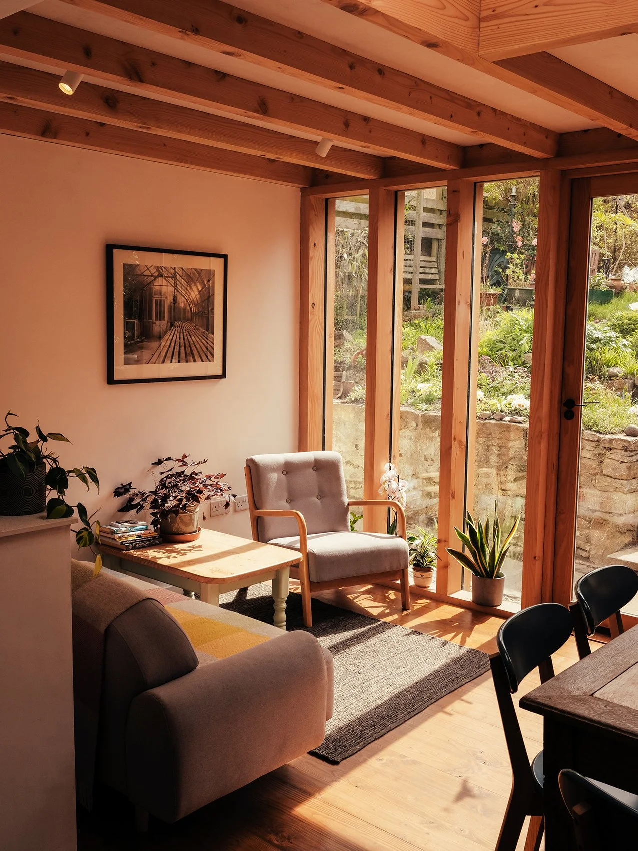 A cozy living room with a beige sofa and a white armchair near a large glass door that opens to a garden, with sunlight streaming in.