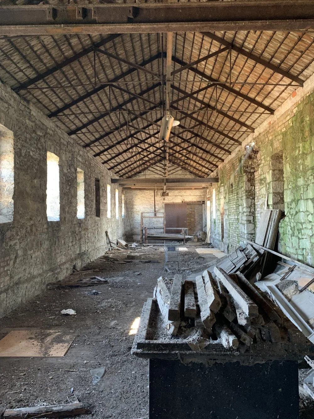 Interior of a dilapidated building under construction or renovation, with debris and construction materials scattered on the floor, exposed brick and stone walls, and a wooden roof structure.