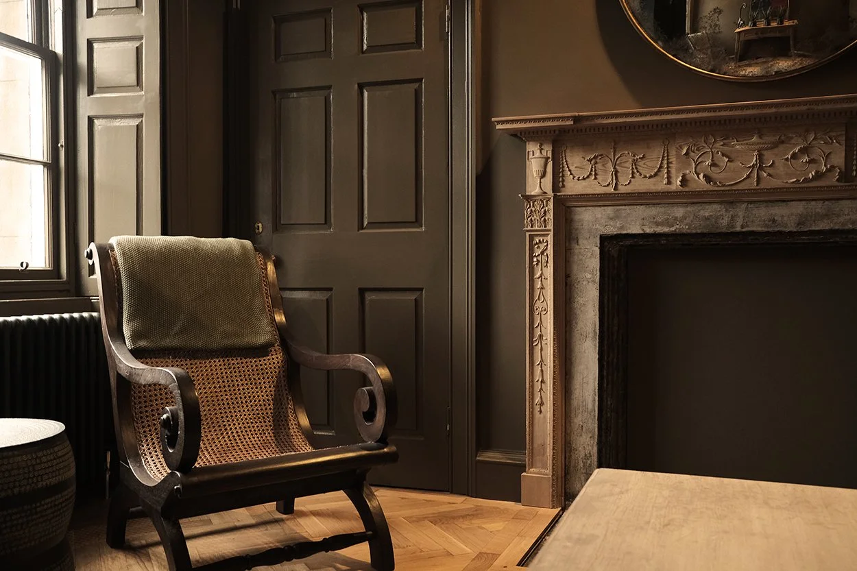A vintage armchair with a cane back and wooden frame in a dimly lit room with a fireplace and dark-colored walls.