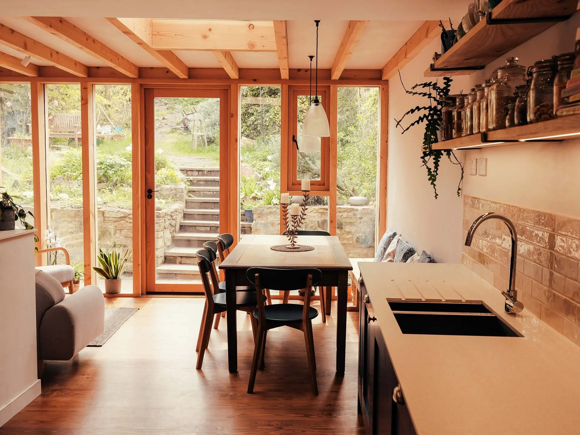 Bright kitchen and dining area with large glass doors leading to outdoor stairs and garden, featuring wooden beams, a dining table with six chairs, a white countertop with a sink, open shelves with jars, a hanging pendant light, and indoor plants.
