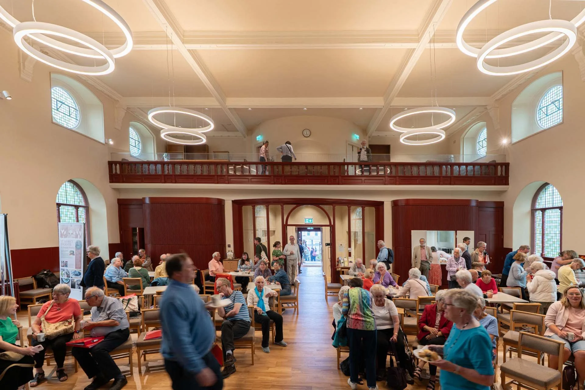 Interior of a large hall filled with elderly people sitting and walking, with wooden accents, high windows, modern circular light fixtures, and a balcony with people standing.