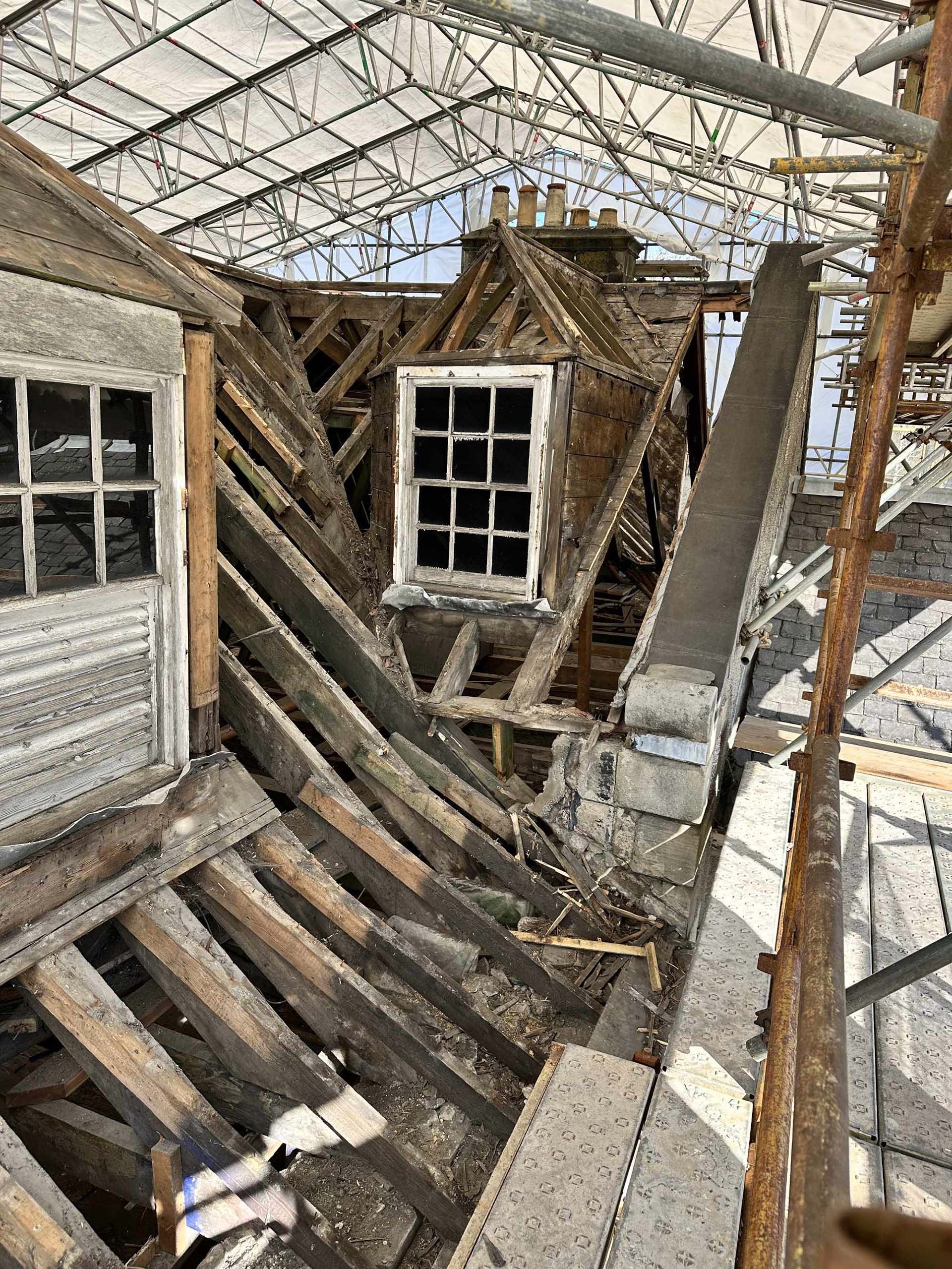 A partially demolished or damaged roof with exposed wooden beams, a small bay window, and scaffolding surrounding the structure.