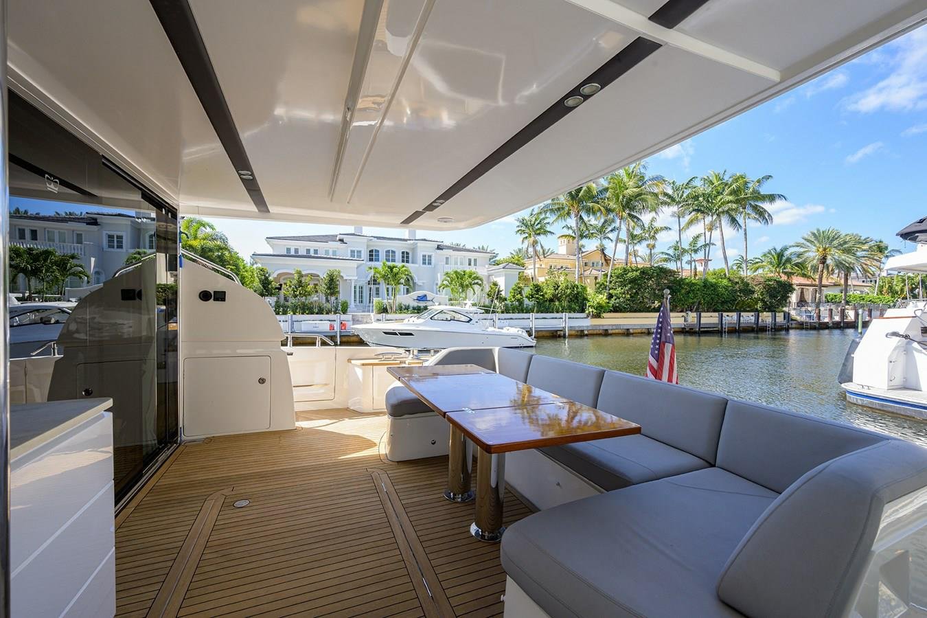 View of a yacht deck with outdoor seating, a wooden table, and a marina with yachts, palm trees, and waterfront houses in the background.