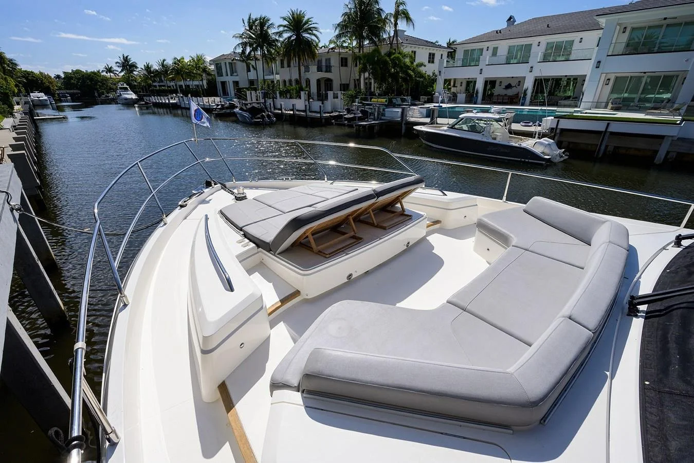 View of a luxury yacht deck with outdoor seating and a sunbed, docked at a marina with water, boats, and modern waterfront houses in the background.
