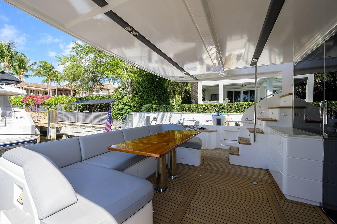Interior view of a yacht's outdoor seating area with a wooden table, white cushioned seats, stairs, and a kitchenette, with boats and trees visible outside.