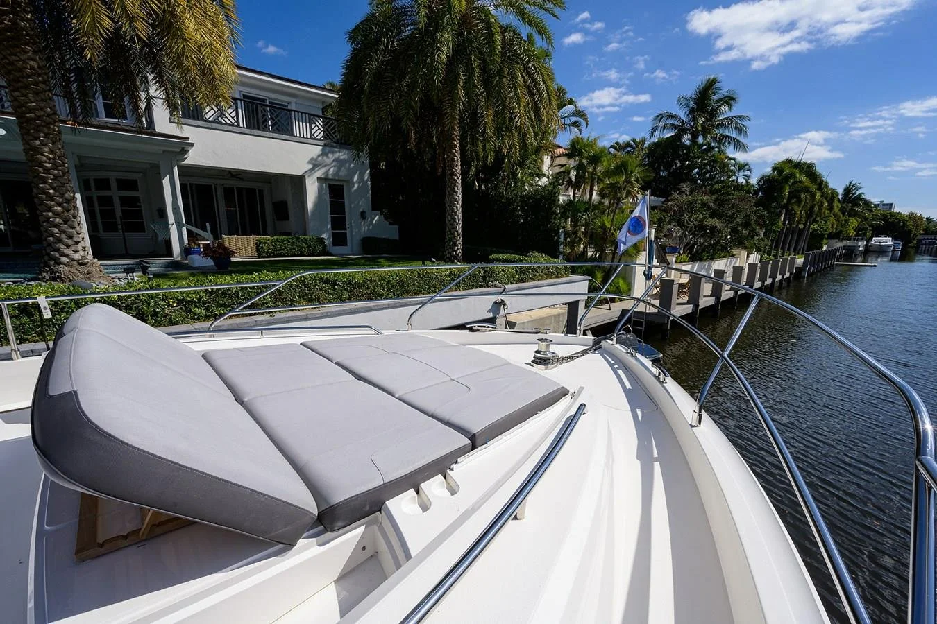 View of a white yacht with a padded sunbathing area on the bow, docked along a canal with houses and palm trees on the waterfront under a partly cloudy sky.
