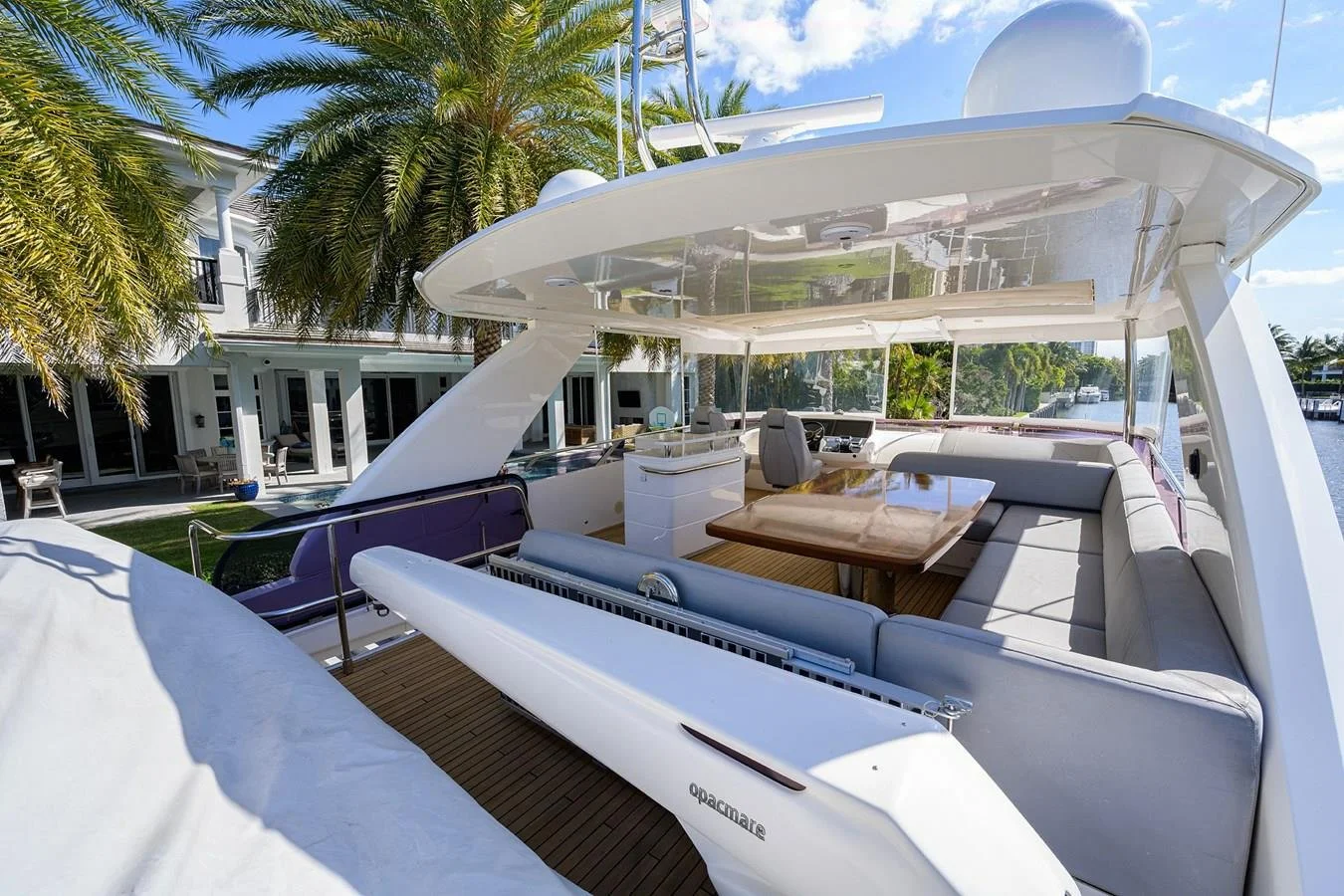 Luxury yacht with seating, a wooden table, and an inflatable boat docked at a canal, with palm trees and a white house in the background.