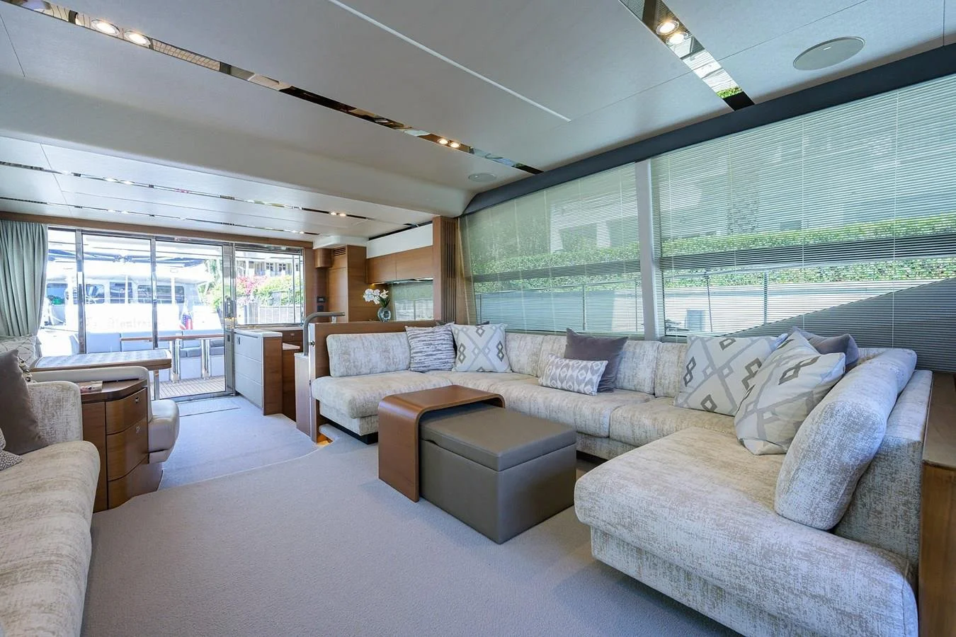 Modern living room with a large L-shaped beige sofa with patterned cushions, large windows with blinds, and glass sliding doors opening to an outdoor patio with a bus outside.