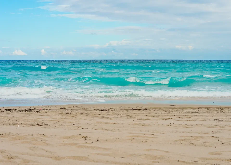View of a sandy beach with turquoise ocean waves and a partly cloudy sky.