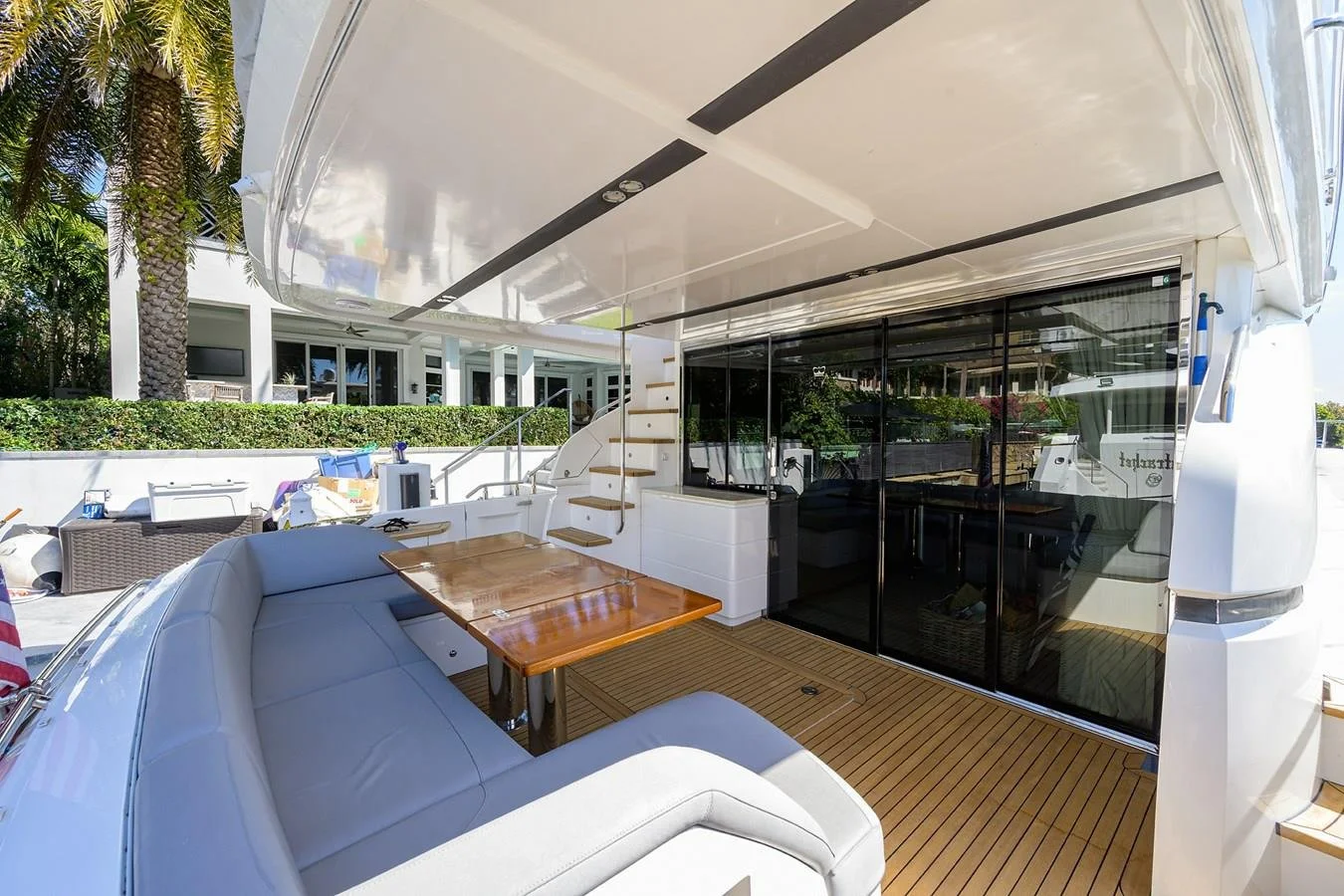 View of the outdoor deck area of a yacht, featuring a white cushioned U-shaped seating area around a wooden table, with stairs and a sliding glass door leading inside, and lush plants in the background.