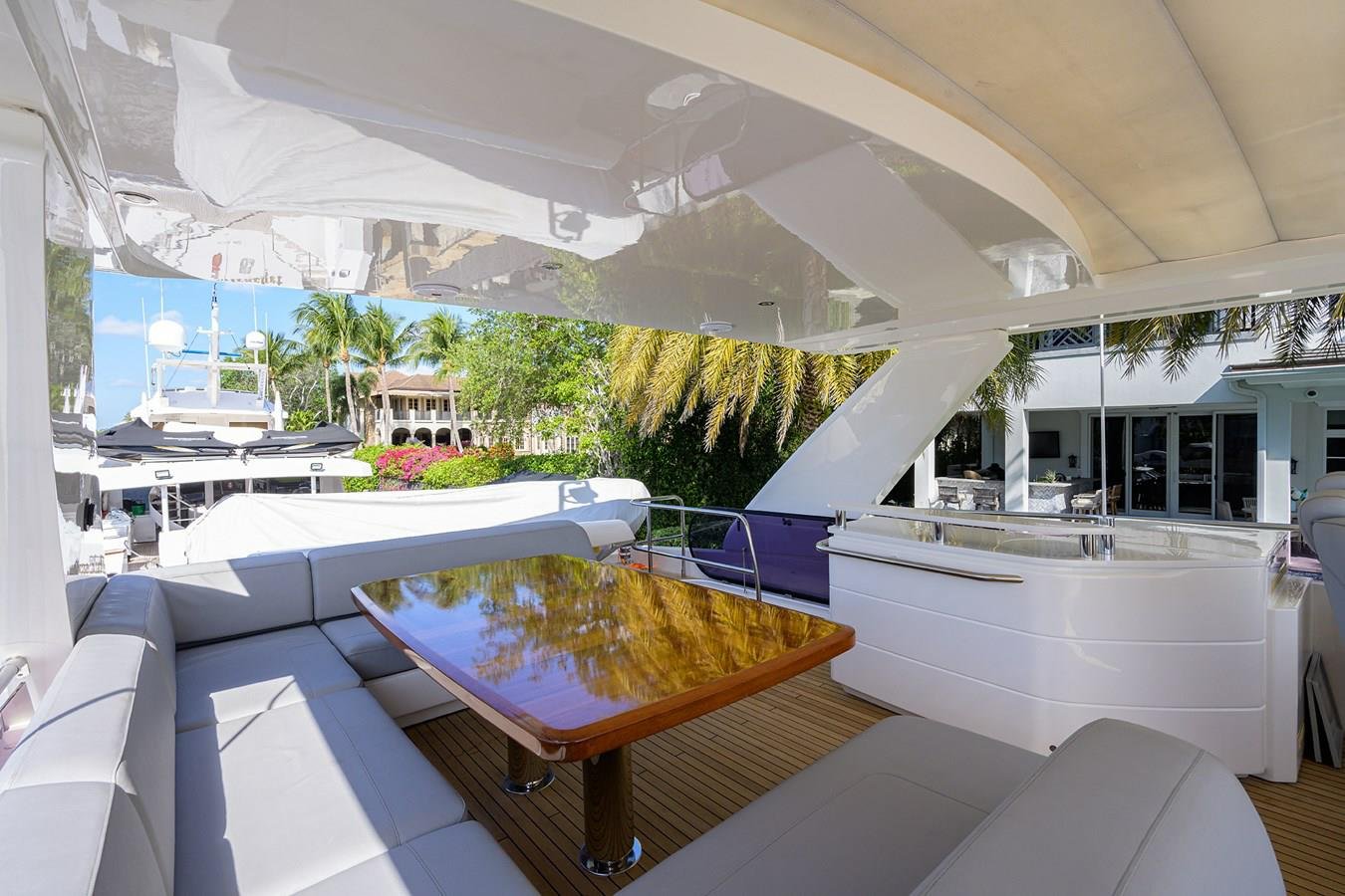 Luxury yacht deck with white seating, a polished wooden table, tropical palm trees, and neighboring yachts and buildings visible in the background.