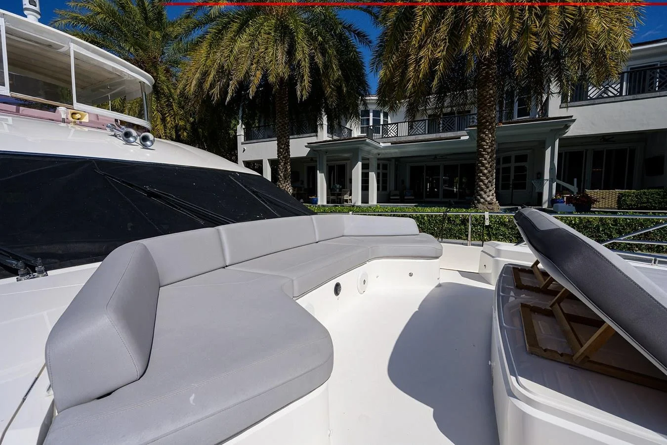 A yacht with gray cushioned seating, a black sunroof, and palm trees in front of a white house with multiple balconies.