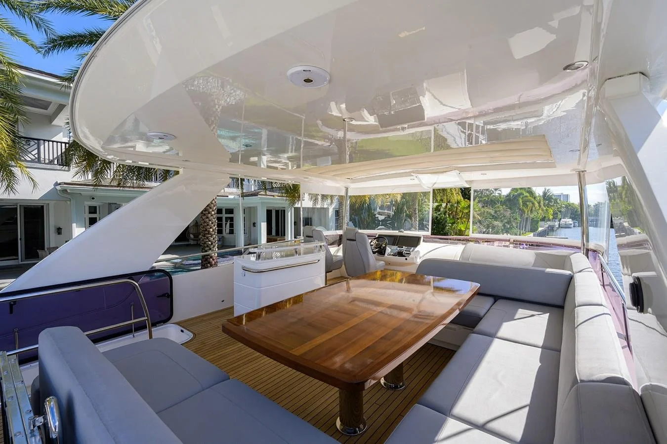 Photo of the interior of a luxury yacht with a wooden table and white cushioned seating, overlooking a marina with palm trees and waterfront houses.