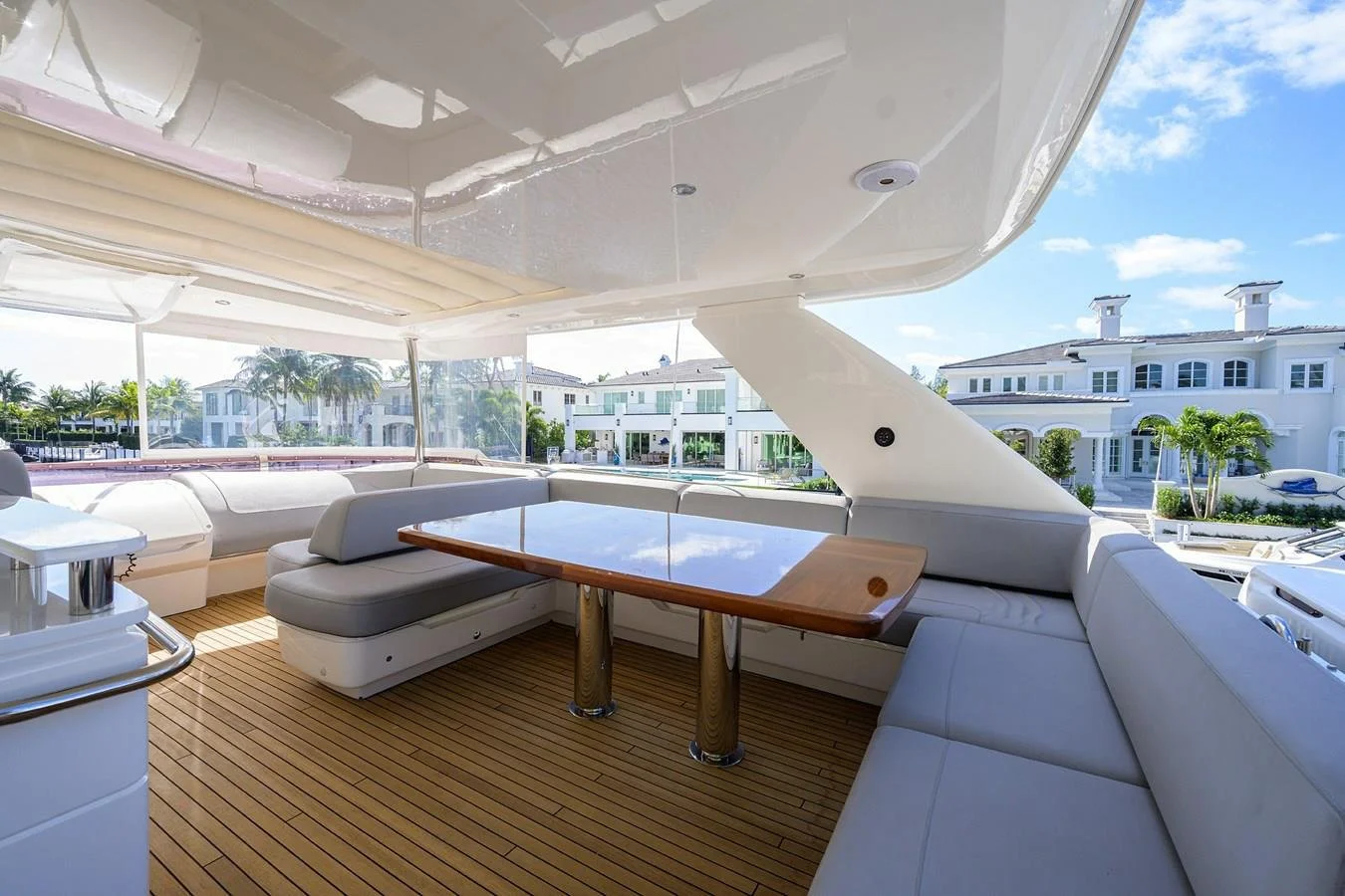 View of the deck area on a yacht with white cushioned seating, a wooden table, and a view of a residential neighborhood with white houses and palm trees.