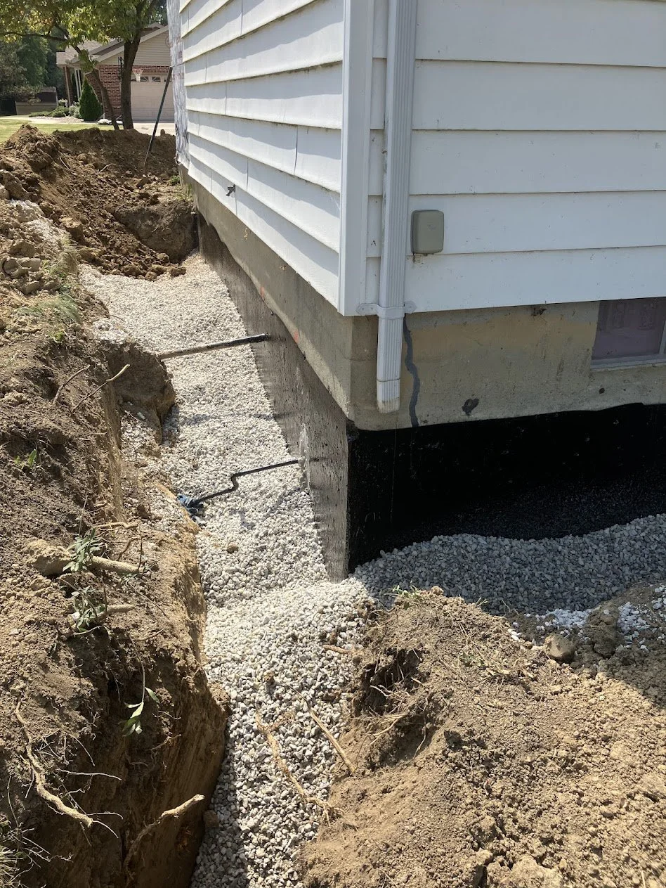 Construction site showing the foundation of a house with gravel and concrete, with exposed dirt and soil, and a white house exterior with siding.