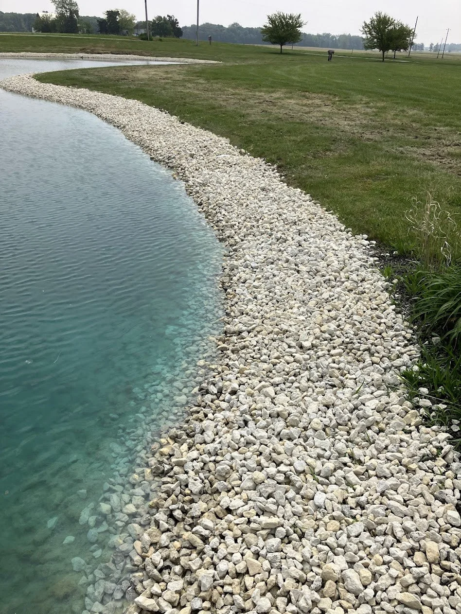 Gravel shoreline along a body of water with green grass and trees in the background.