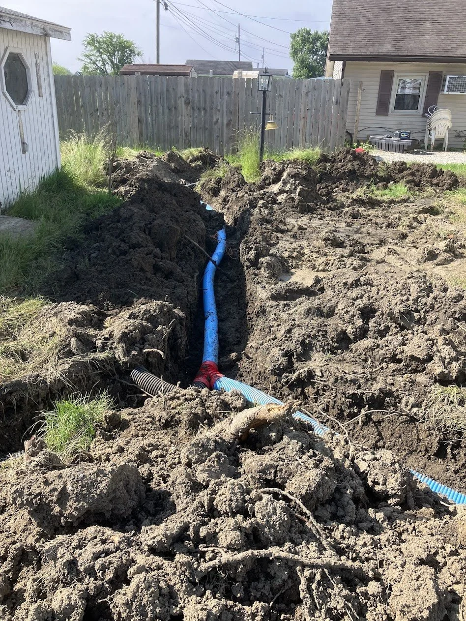 A deep trench in a backyard with underground pipes, including a blue pipe and a black corrugated pipe, surrounded by dirt and grass, with a wooden fence and a house in the background.