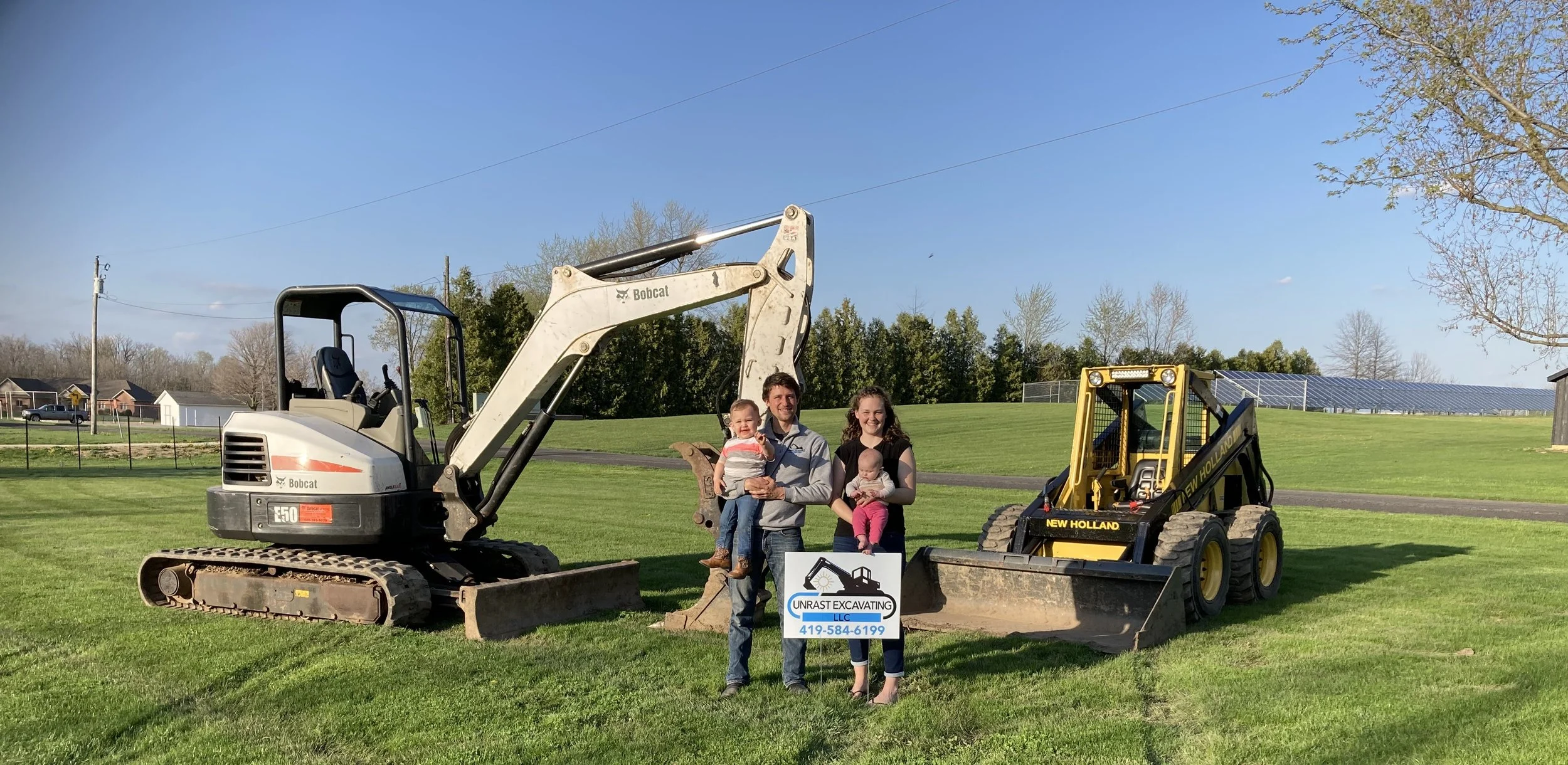 Family standing between two small excavators holding a sign for Unrast Excavating LLC, on a grassy field with trees and solar panels in the background.