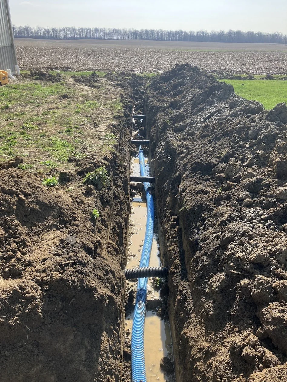 A trench dug in the ground with blue piping and black corrugated conduit, possibly for underground utility installation, amid a rural landscape with fields and trees in the background.