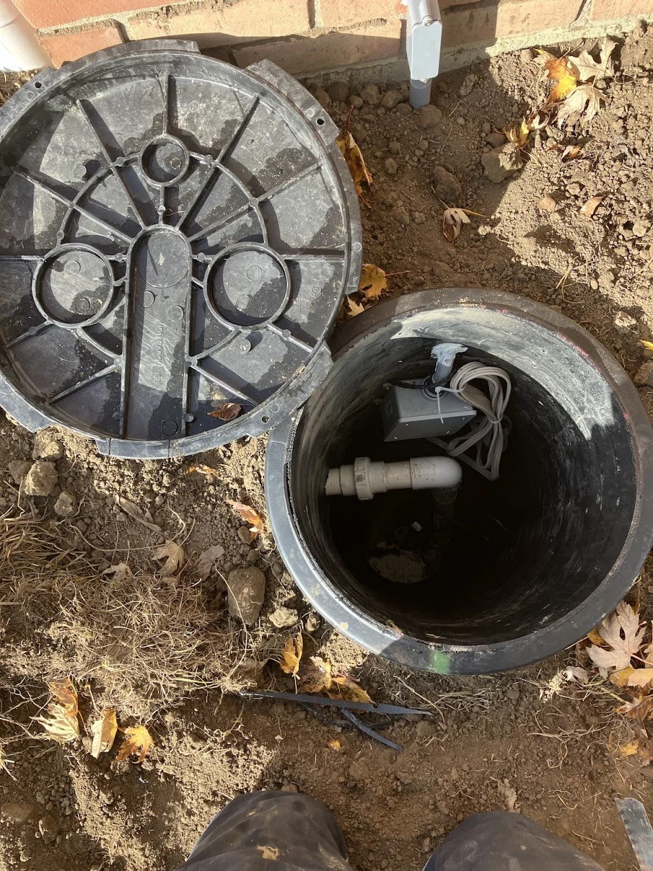 An underground utility box with a round cover removed, revealing electrical components and wiring inside, surrounded by soil and fallen leaves.