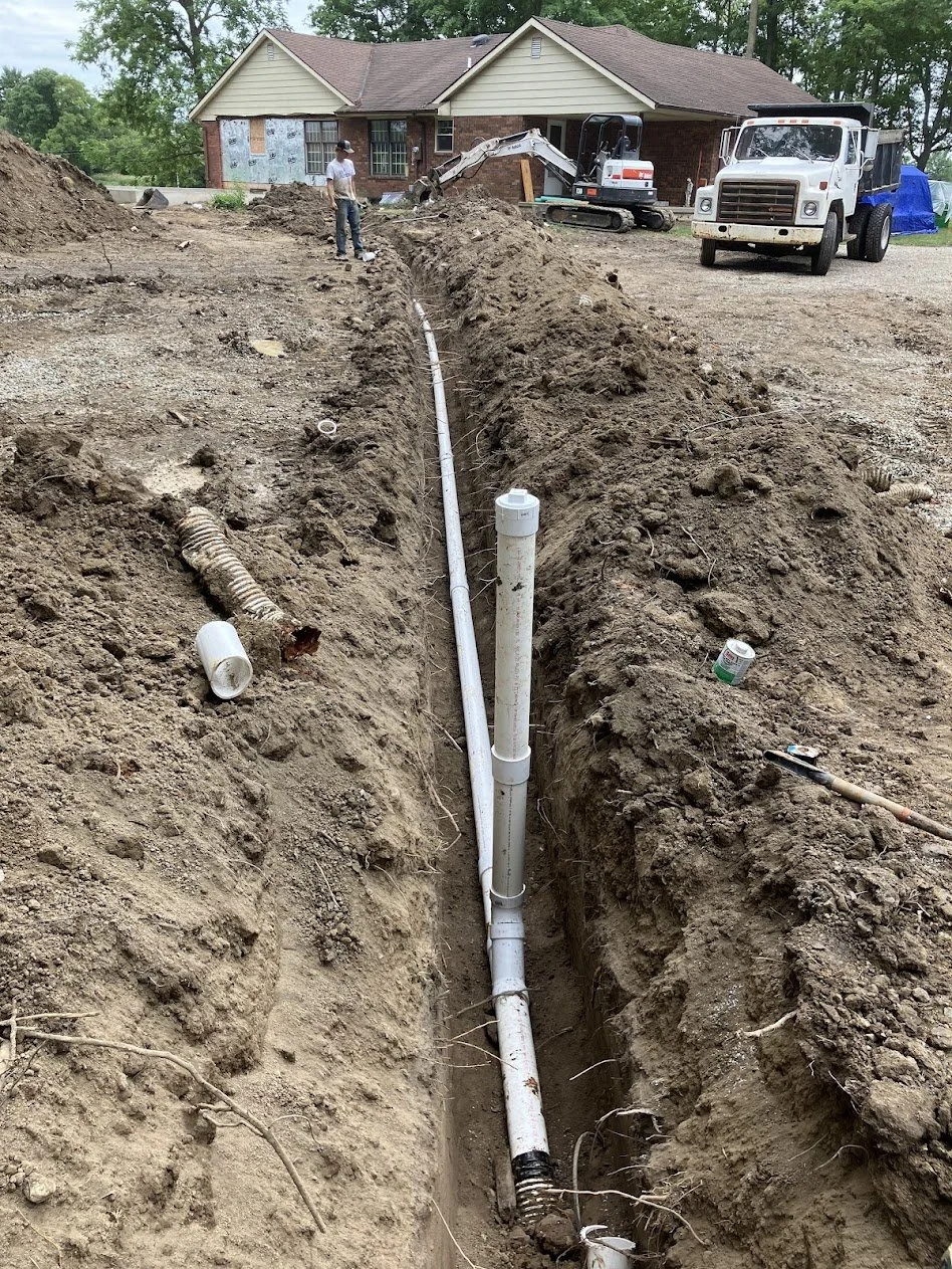 Excavation site with a trench for underground utility pipes, construction tools, and a worker in front of a house undergoing renovation or construction.