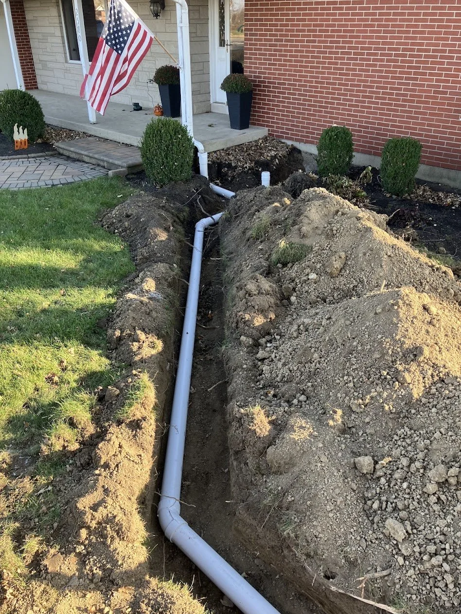 A trench with a new PVC pipe being installed outside a house, with a American flag, small shrubs, and a brick and siding house in the background.