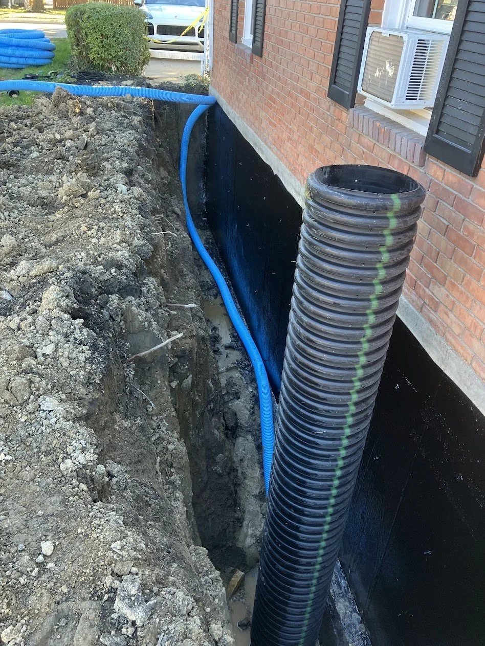 Construction site showing excavation along the side of a brick house, with black waterproofing and blue corrugated drainage pipes installed near the foundation.
