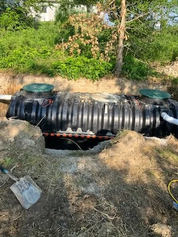 Underground black corrugated septic tank with visible plumbing, partially buried in dirt with grass and trees around it.