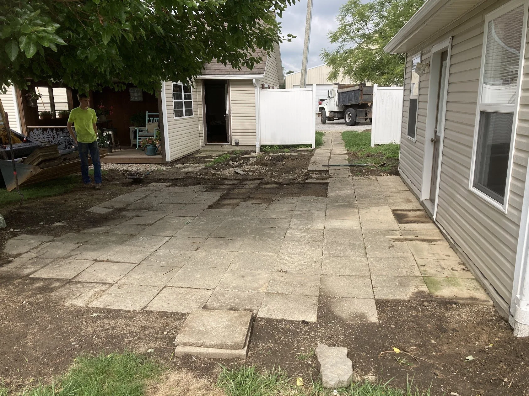 Backyard with an ongoing patio renovation, showing a partially removed pavedstone patio, a person in a bright yellow shirt, narrow concrete walkway, small shed, and a white fence.
