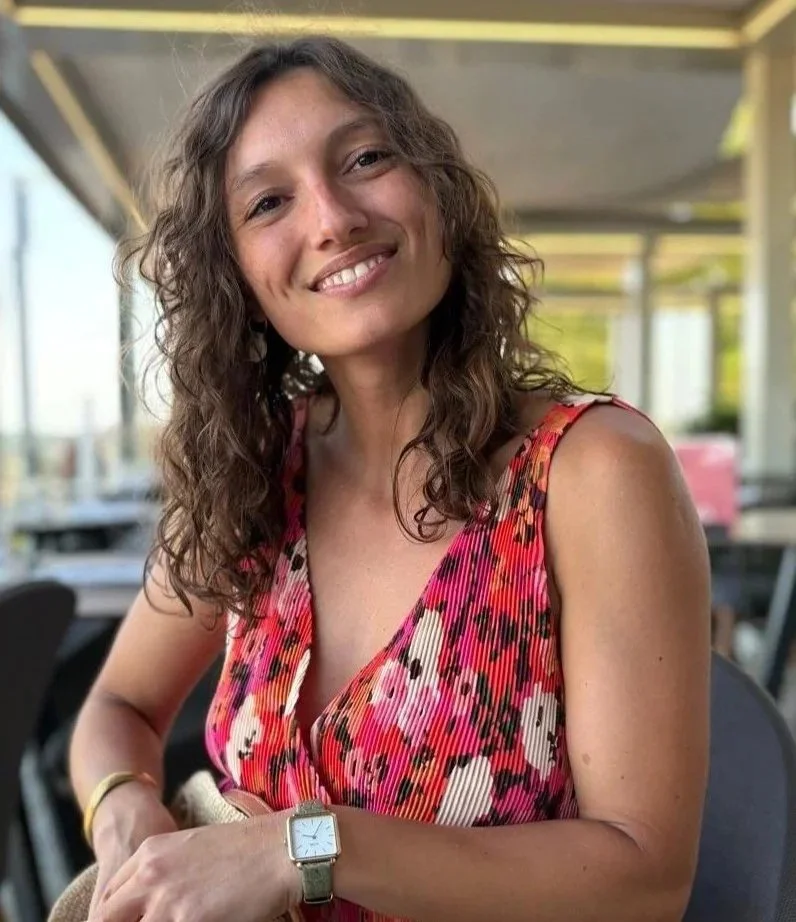Une femme souriante avec des cheveux ondulés, portant une robe à motifs rouges et roses, assise dans un restaurant ou un café avec un décor moderne et de grandes fenêtres.