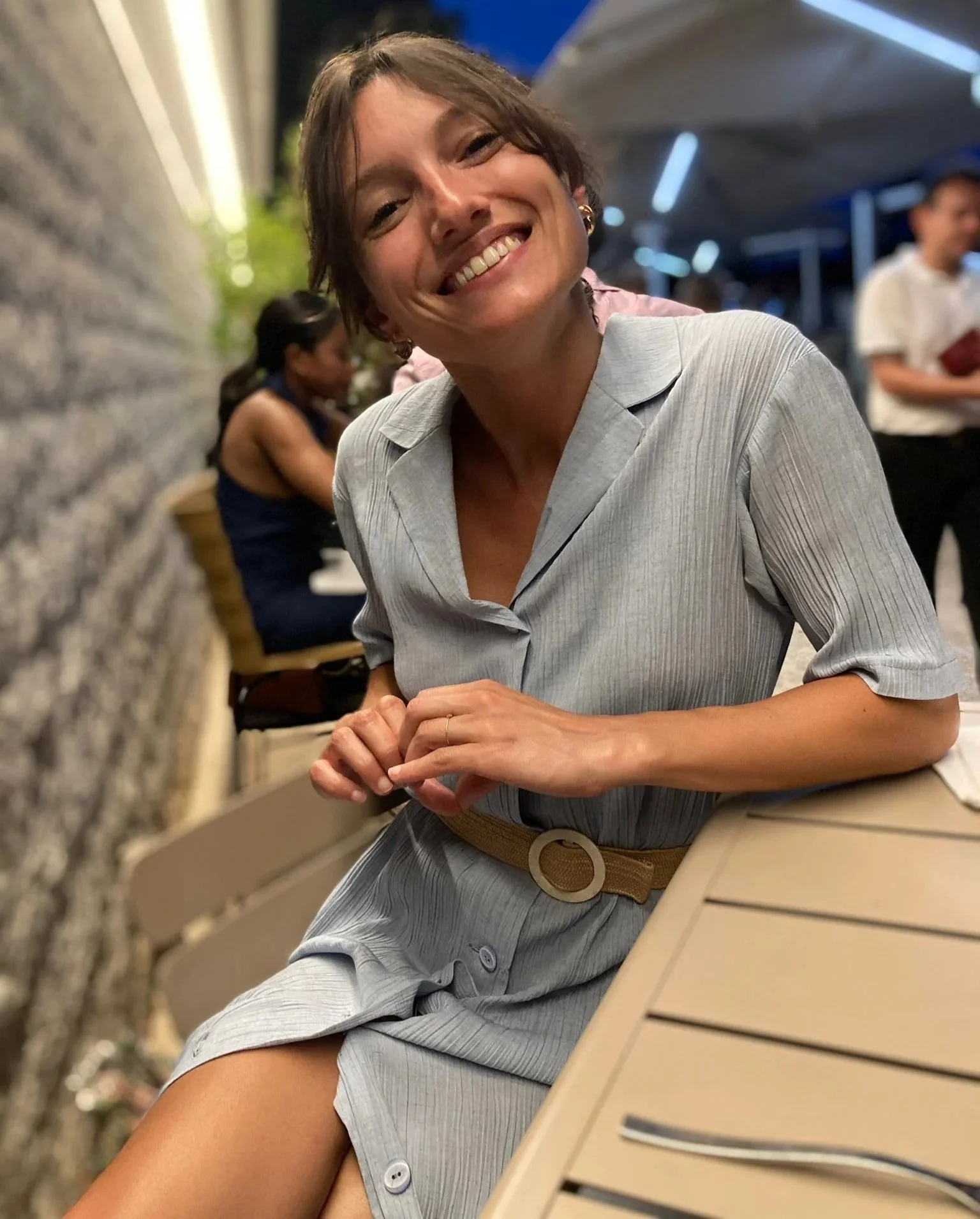 Une femme avec une robe grise assise à une table en extérieur, souriant à la caméra, avec d'autres personnes en arrière-plan dans un environnement de restaurant ou café.