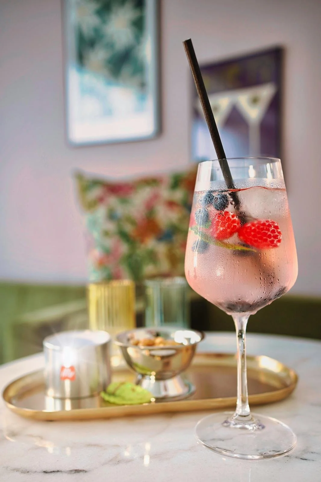 Ein großes Glashalter mit einem rosa Getränk, gefüllt mit Beeren wie Himbeeren, Heidelbeeren und Brombeeren, mit einem schwarzen Strohhalm auf einem Tisch, daneben eine goldene Tablett mit kleinen Schälchen und einem grünen Macaron, im Hintergrund zw