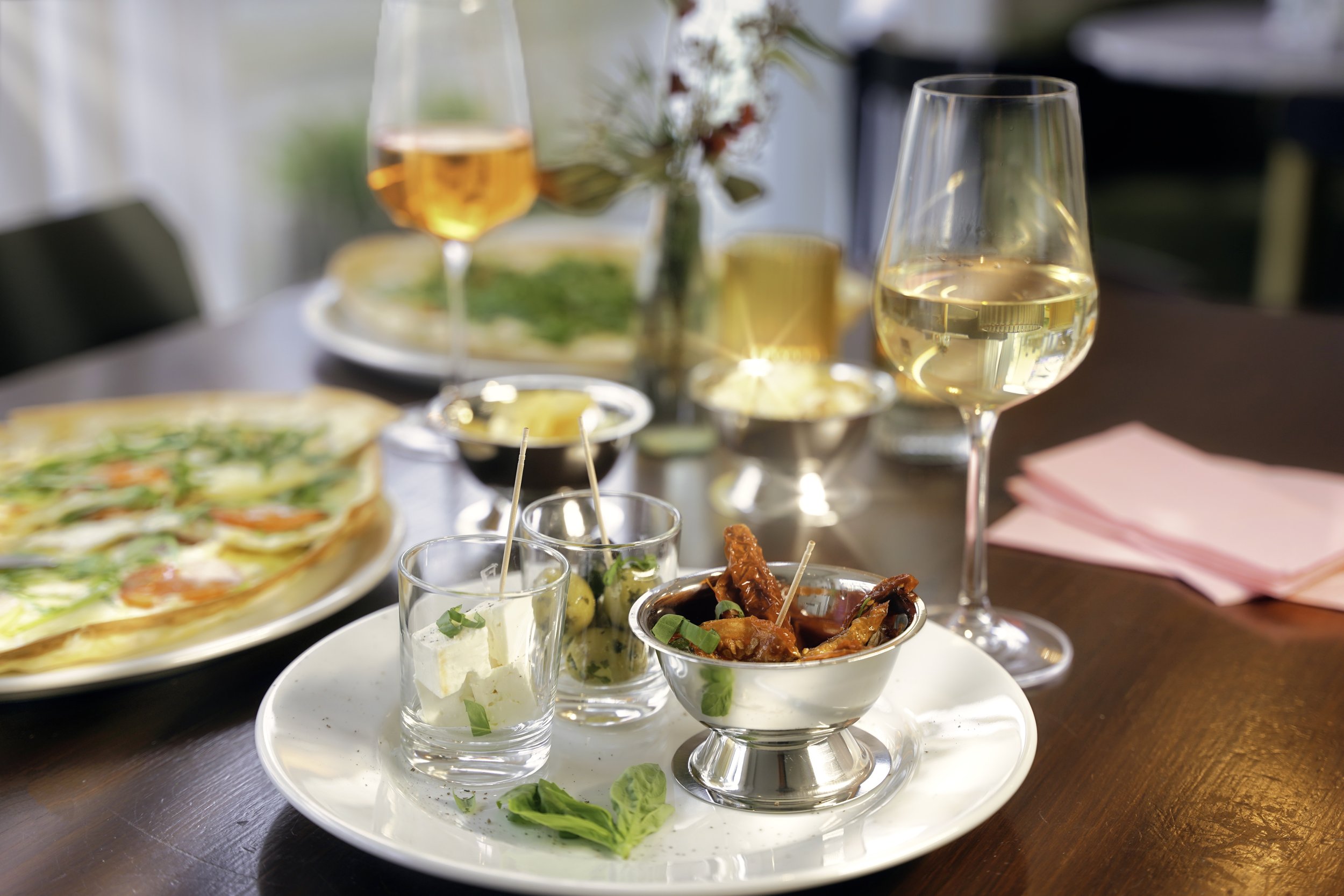 Vorspeisenteller mit verschiedenen kleinen Gerichten, zwei Gläser Roséwein, Pizza, Pickles, Oliven, und weitere Speisen auf einem Tisch bei einem Essen
