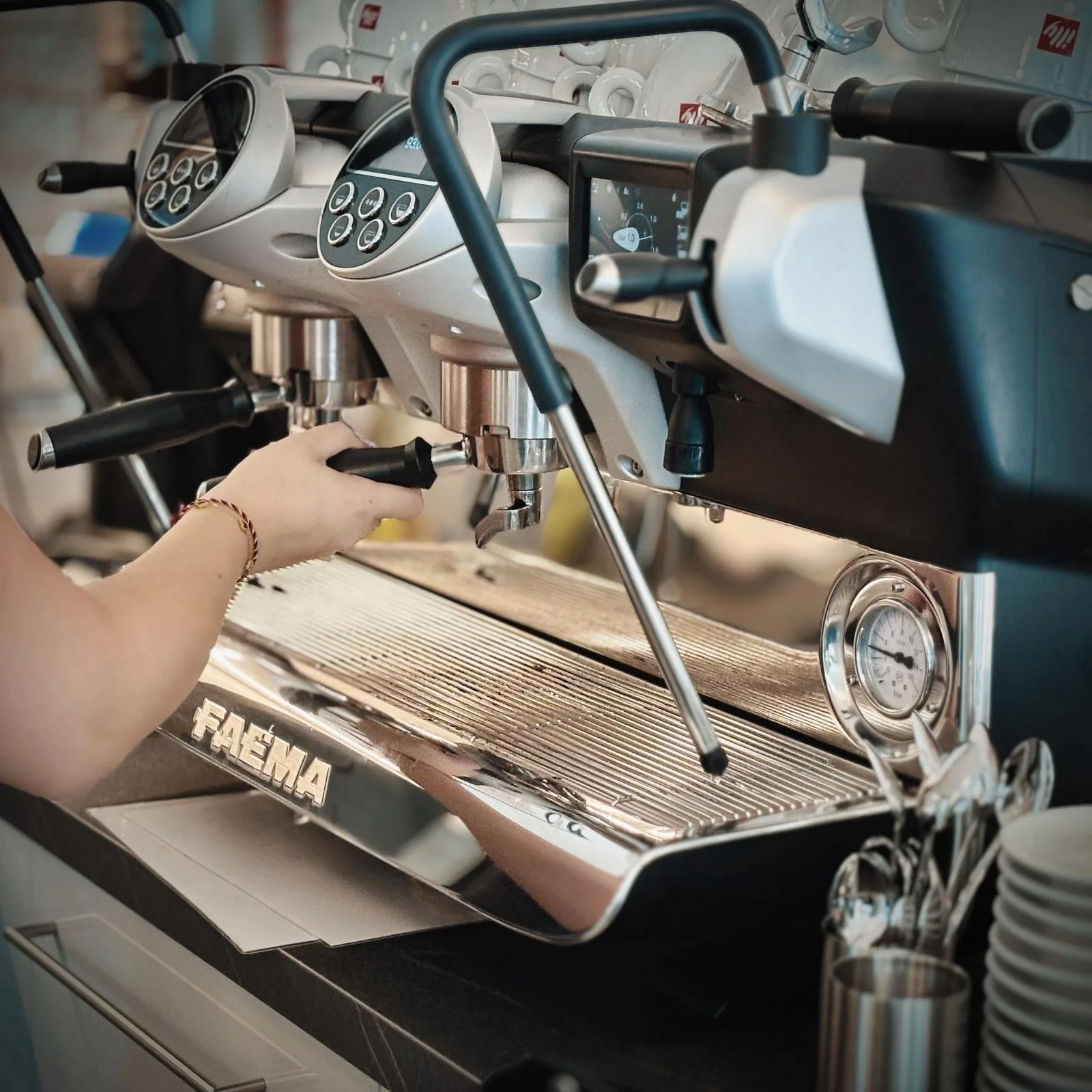 Eine Espressomaschine auf einem Tresen, jemand bereitet Espresso zu, mit einem Stabmixer oder Düse. Es gibt einen Druckmesser und verschiedene Tasten auf der Maschine. Im Hintergrund befinden sich weiße Tassen.