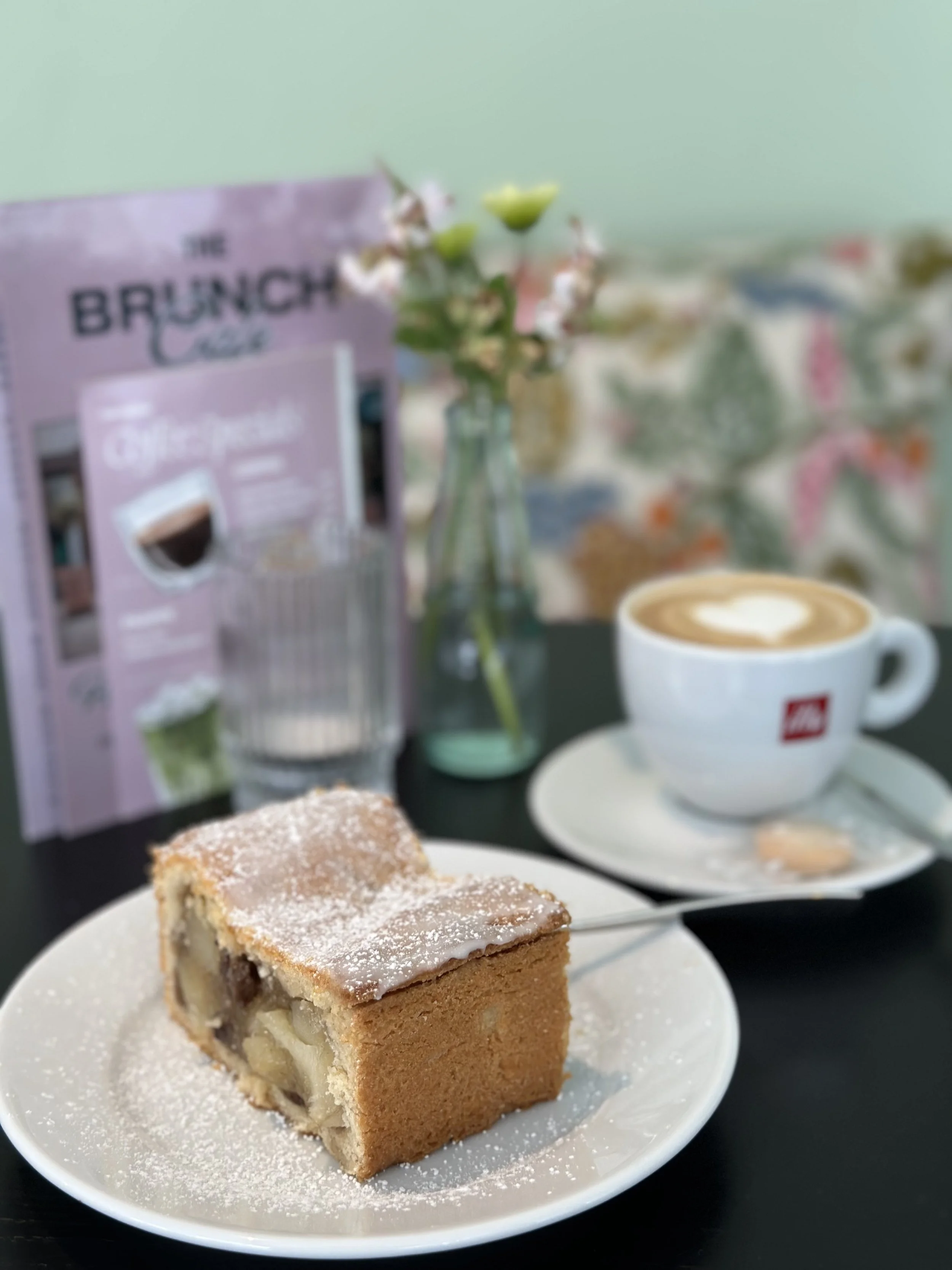 Ein Teller mit einem Stück Apfelkuchen, eine Tasse Kaffee mit Latte Art und eine Vase mit kleinen Blumen, im Hintergrund eine Verpackung des Kaffeekapselherstellers.