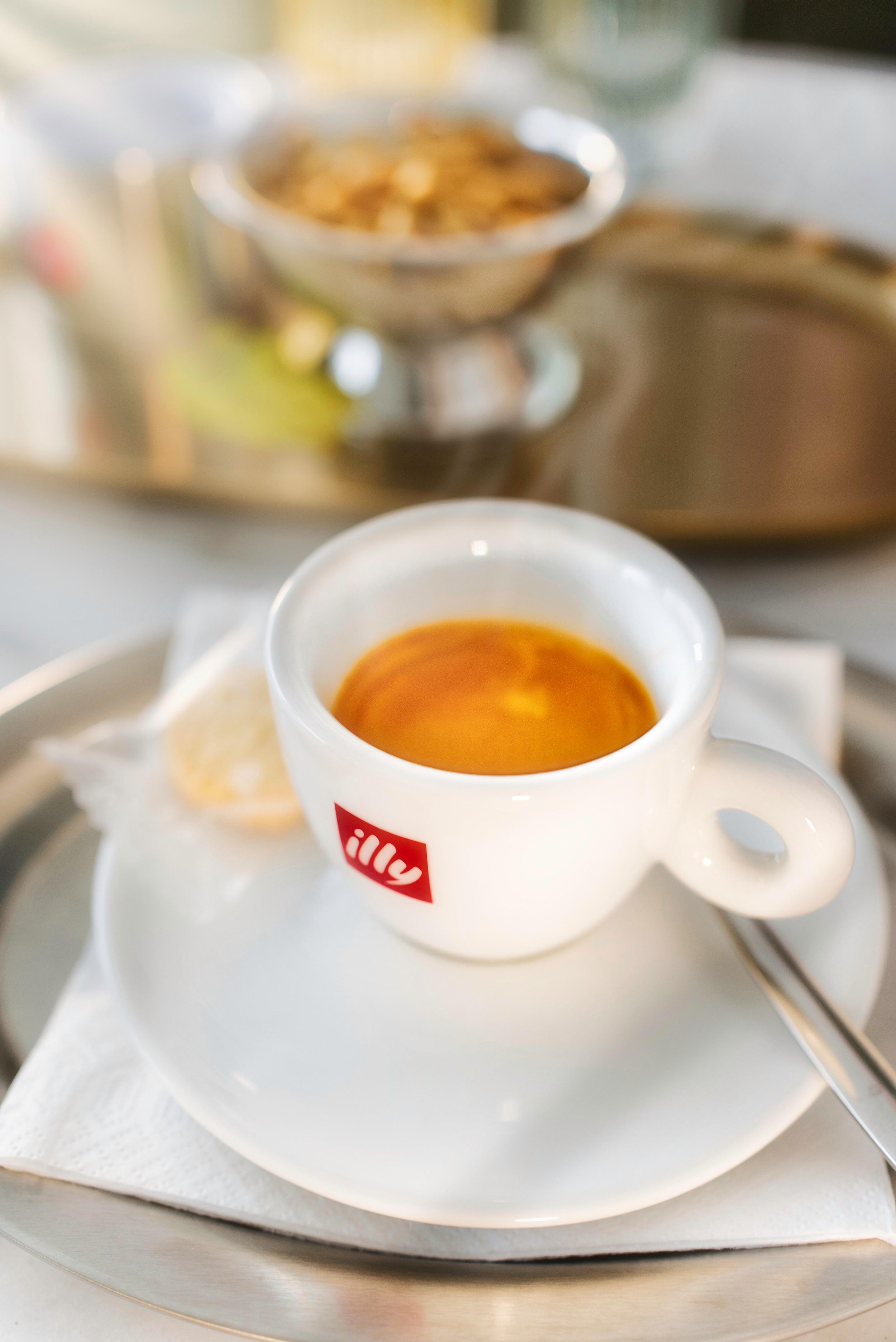 Eine Tasse mit Espresso auf einem weißen Teller, daneben ein Löffel, auf einem Tablett mit Papierserviette, im Hintergrund eine Schüssel mit Keksen.