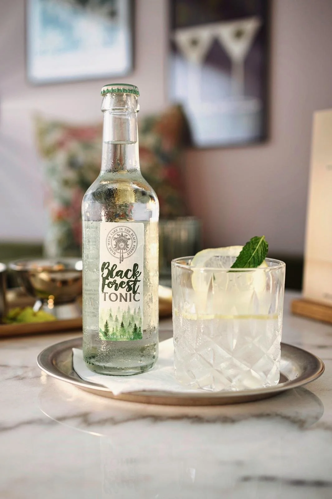 Eine Flasche Black Forest Tonic und ein Glas Gin mit Zitrone und Minze auf einem silbernen Teller auf einem weißen Marmortisch.