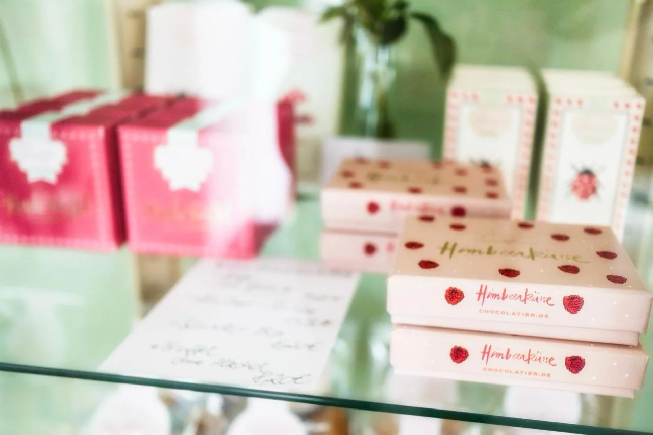 Verpackte Himbeerküsse auf einem Glastisch, im Hintergrund pinke Verpackungen und eine Vase mit grünen Blättern.