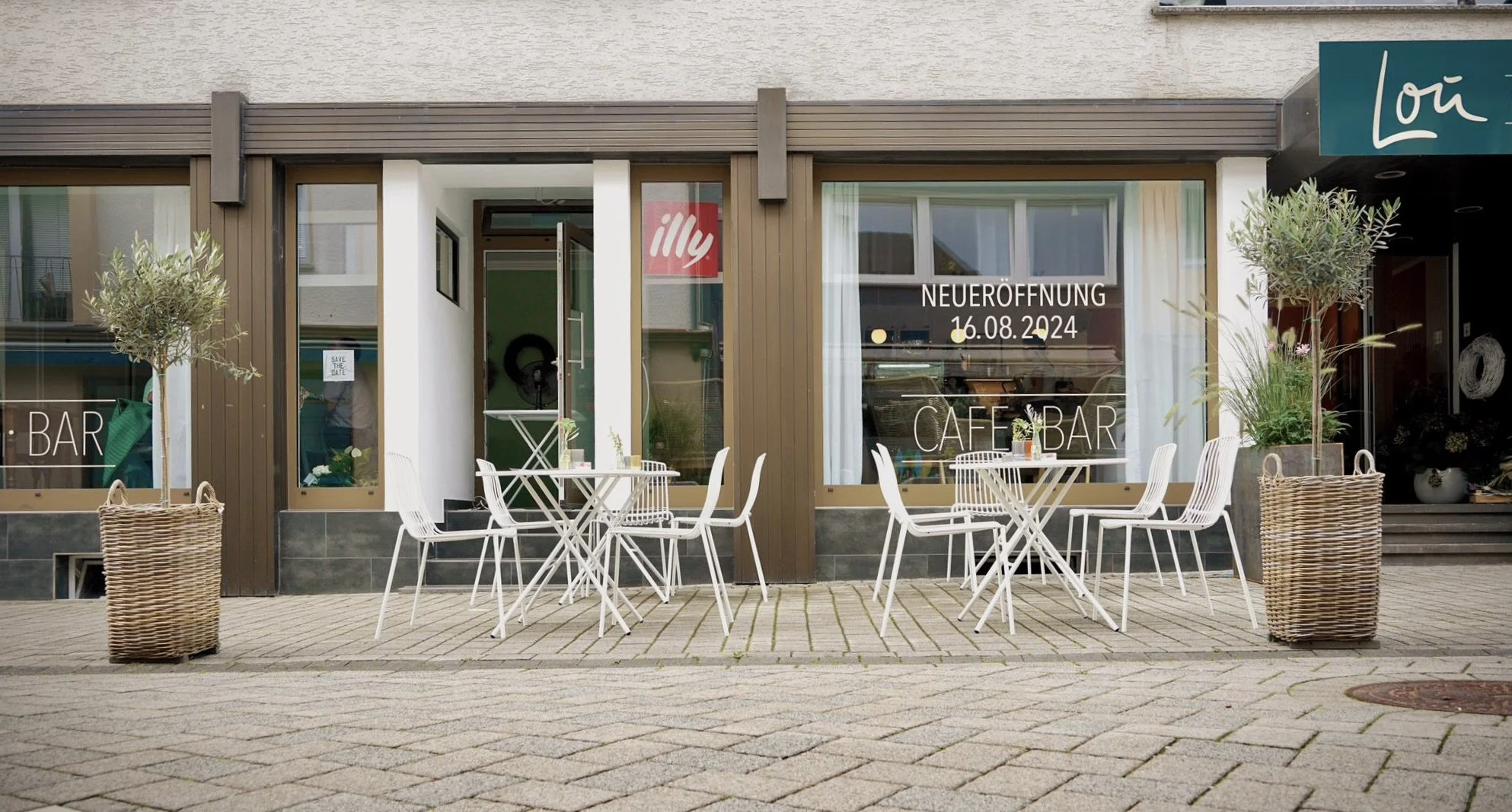 Außenansicht eines Cafés mit weißen Tischen und Stühlen auf dem gepflasterten Gehweg, große Pflanzen in Körben an den Fenstern, Hinweis auf Neueröffnung am 16.08.2024, Schilder für Bar und Café-Bar.