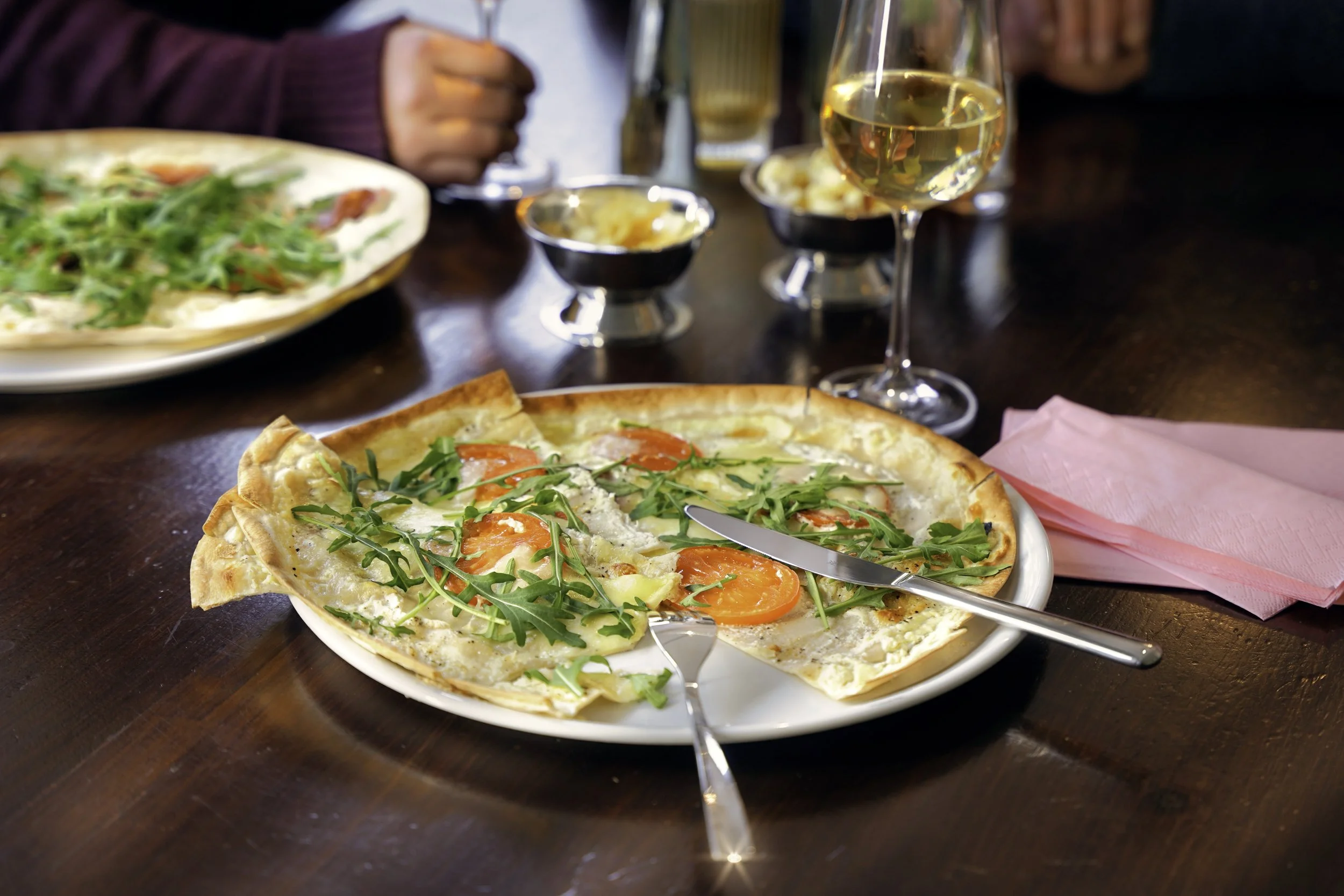 Flammkuchen mit Tomaten, Rucola und Käse auf einem weißen Teller, in der Nähe ein Glas Weißwein, im Hintergrund weitere Speisen und Getränke auf einem dunklen Holztisch.