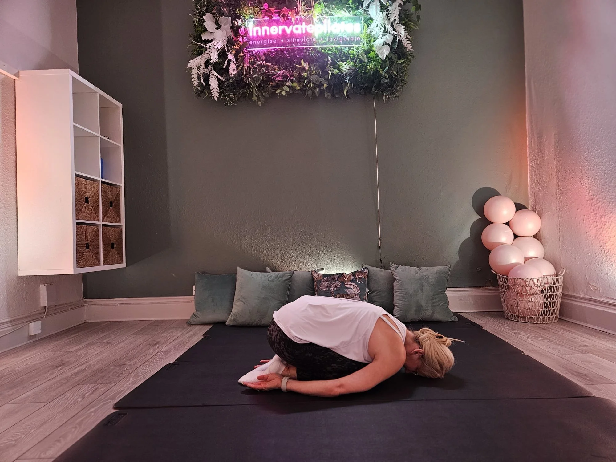 A woman practicing yoga in a child's pose on a black yoga mat in a room with wooden floors and gray walls. Behind her are large pillows, a decorative basket with pink balloons, a bookshelf, and a wall decoration with a neon sign that reads 'innervate