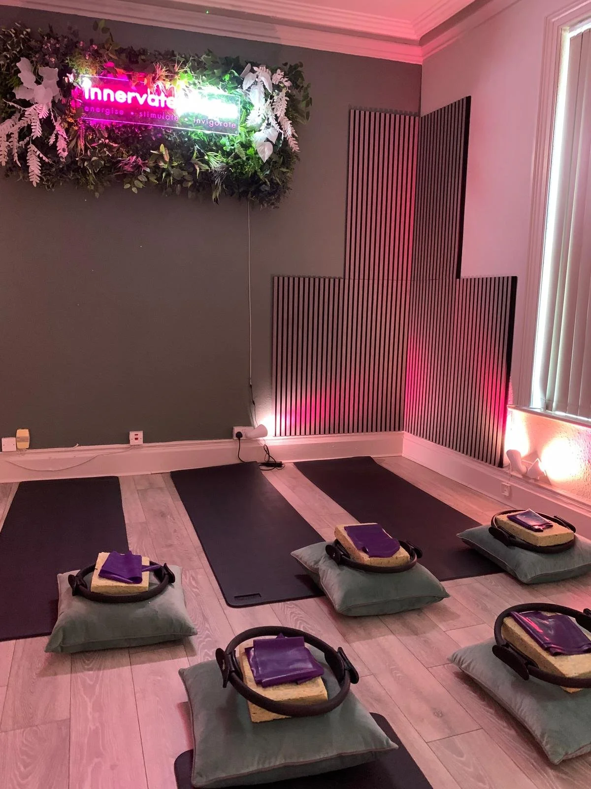 A yoga or meditation class setup in a studio with six black mats and green cushions, each with a yoga block and strap, arranged on a wooden floor. The room has a gray wall with decorative black and white panels and a neon sign that reads "Innervale" 
