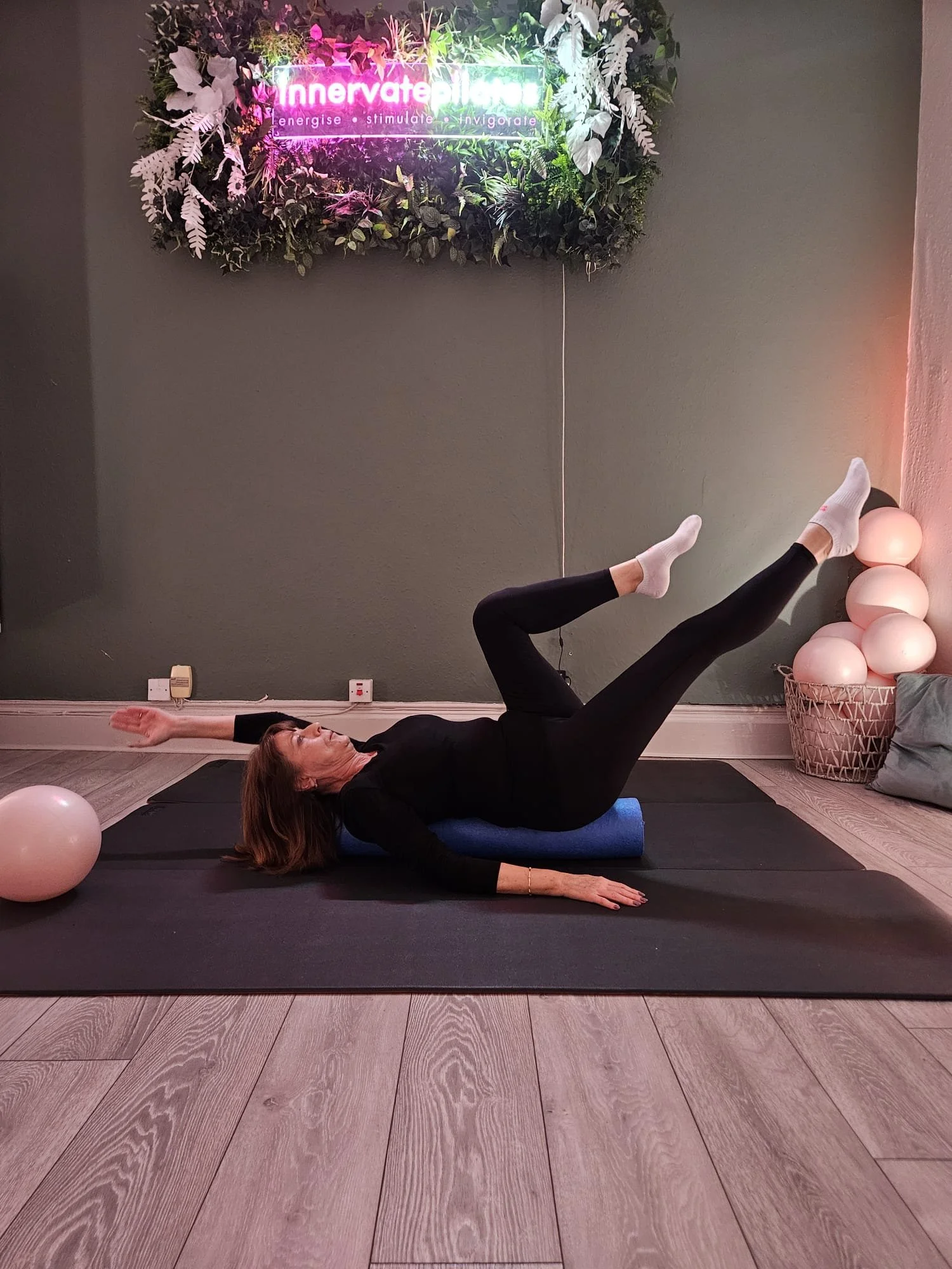 A woman is lying on a yoga mat in a living room, performing a yoga pose with one leg raised and arms extended. She is wearing a black outfit and white socks. The room has wooden flooring, pastel-colored balloons stacked in a basket, and a neon sign o