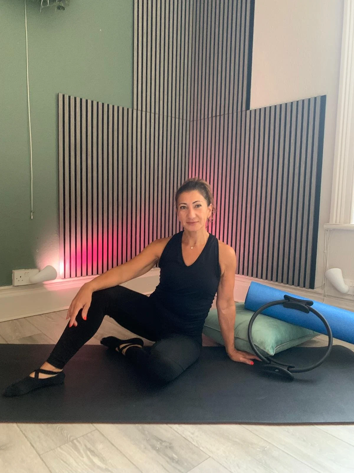 A woman in black workout clothes sitting on a yoga mat in a fitness studio, with exercise equipment.
