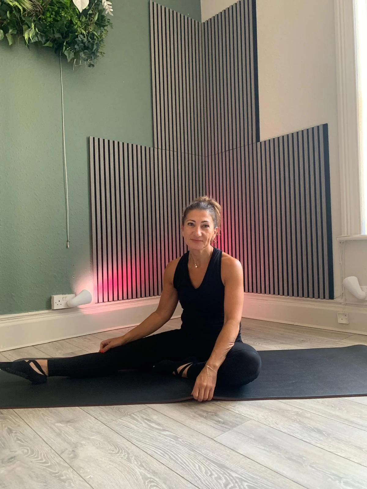 A woman sitting on a yoga mat in a yoga studio with sound panels on the wall and a potted plant hanging from the ceiling.