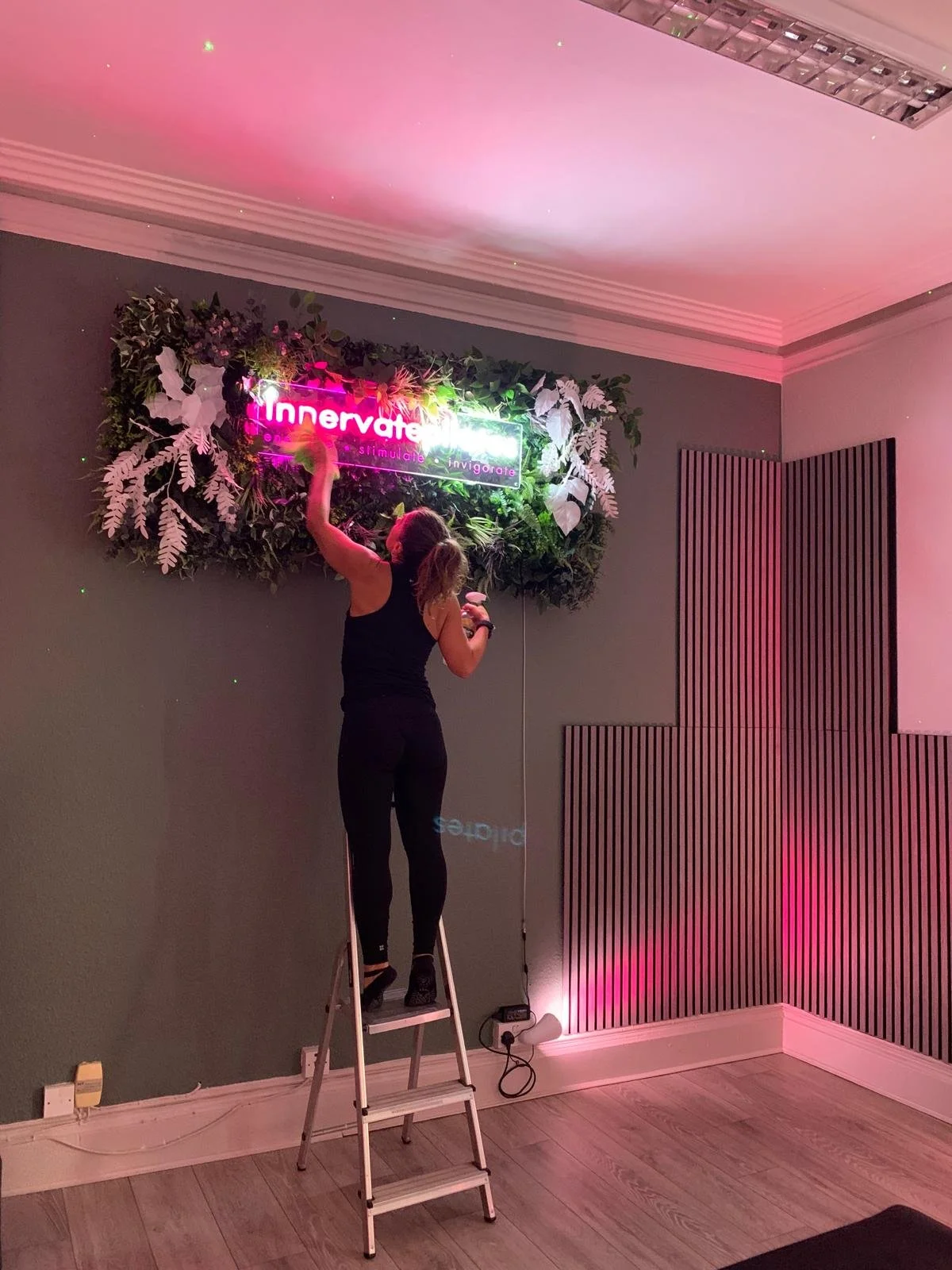 A woman standing on a ladder attaching a neon sign to a wall decorated with artificial greenery and white leaves. The neon sign reads 'Innerve' with words 'stimulate' and 'invigorate' underneath, and the room has pink lighting with wooden wall panels