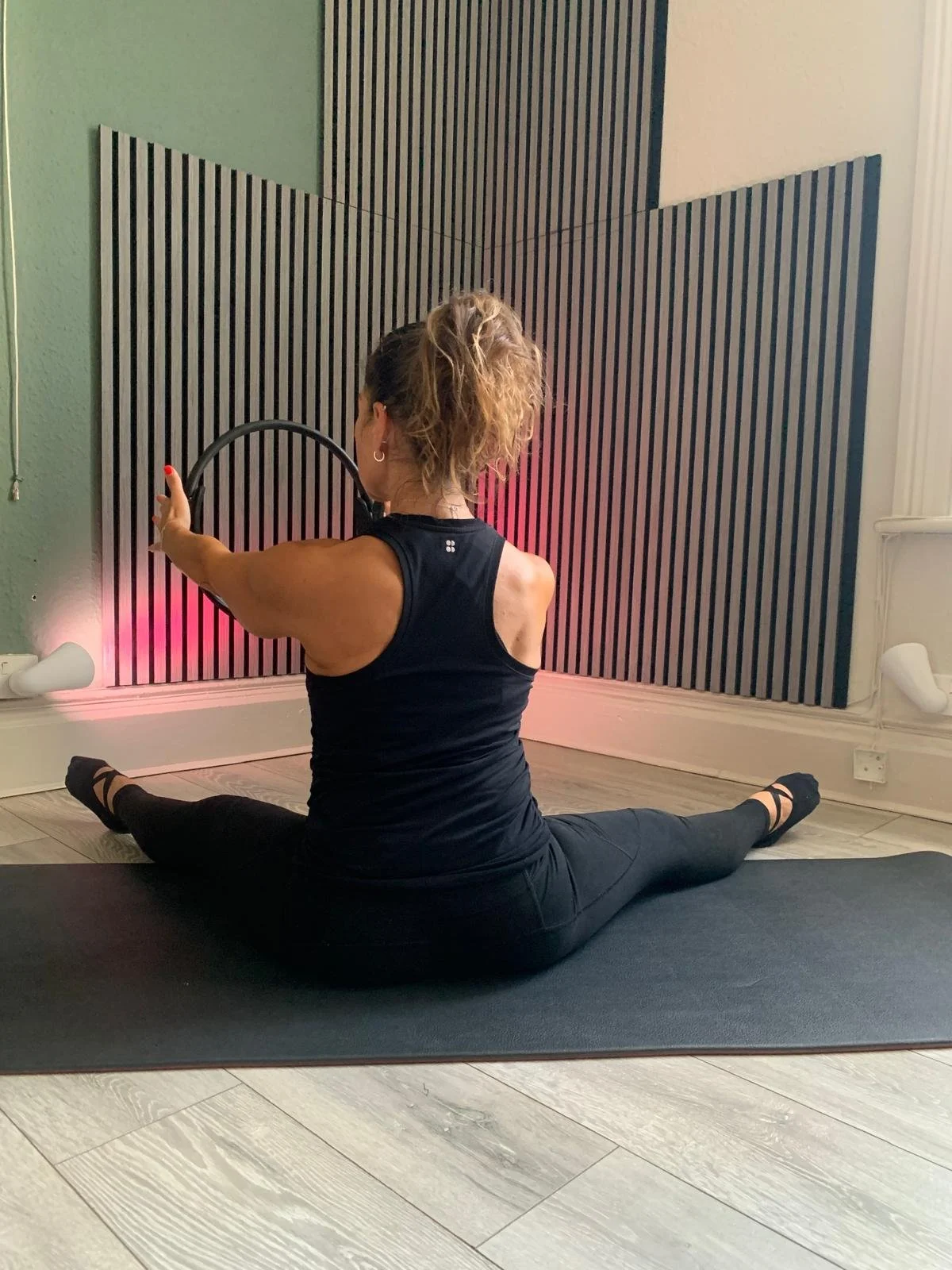 Woman practicing yoga in a split sit stretch, holding a hoop with one arm in a room with black and white acoustic panels and hardwood flooring.
