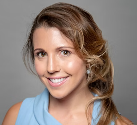 A smiling woman with light brown hair styled in loose waves, wearing a blue sleeveless top and pearl earrings, against a gray background.