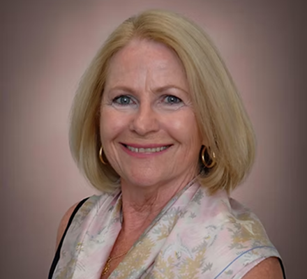 A smiling woman with shoulder-length blonde hair wearing gold hoop earrings and a pastel patterned blouse against a neutral background.